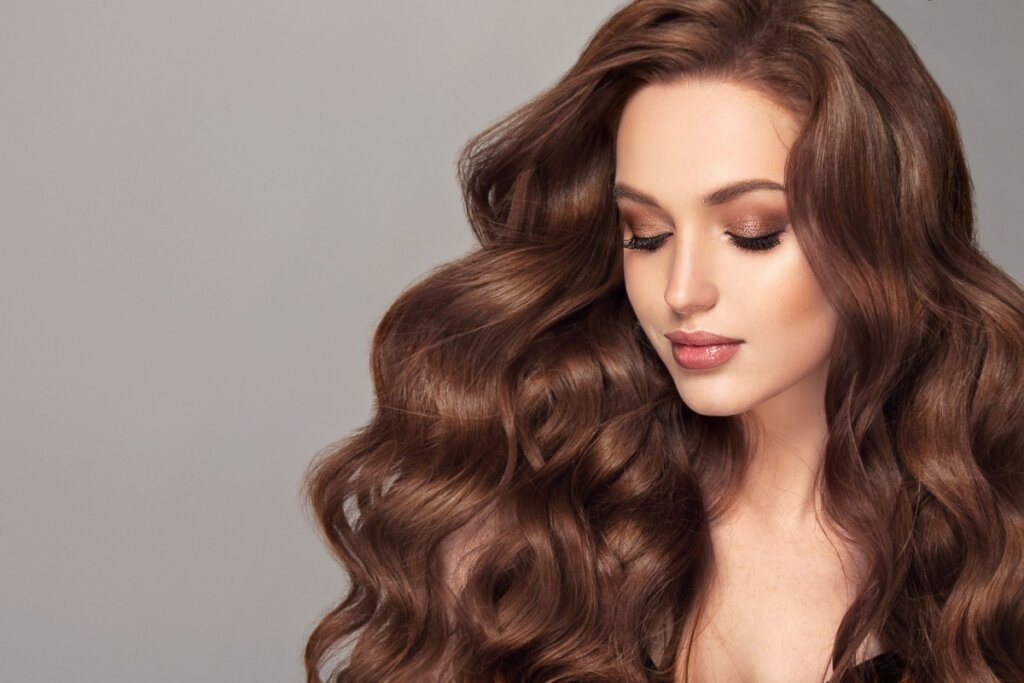Mulher jovem com cabelo longo e ondulado em tom castanho chocolate intenso, com brilho e volume, usando maquiagem suave e olhando para baixo sobre fundo neutro