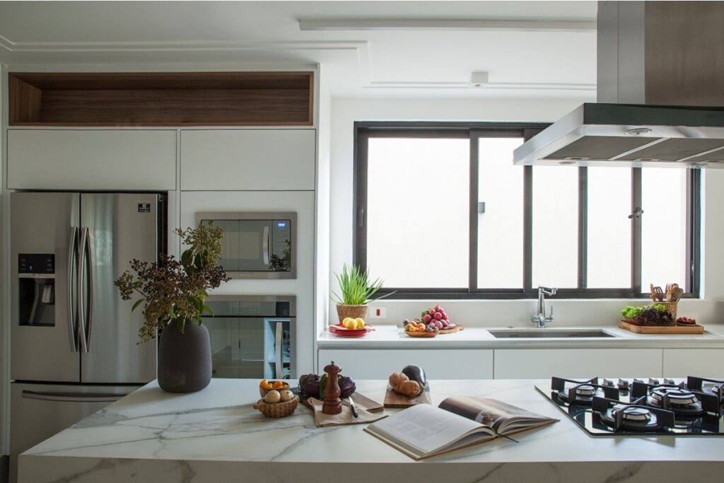 Cozinha ampla com ilha central em mármore branco. Na ilha, há um cooktop e um livro de receitas aberto. Ao fundo, uma bancada com cuba simples de inox posicionada sob uma grande janela de esquadria preta. A marcenaria é branca com nichos amadeirados superiores.