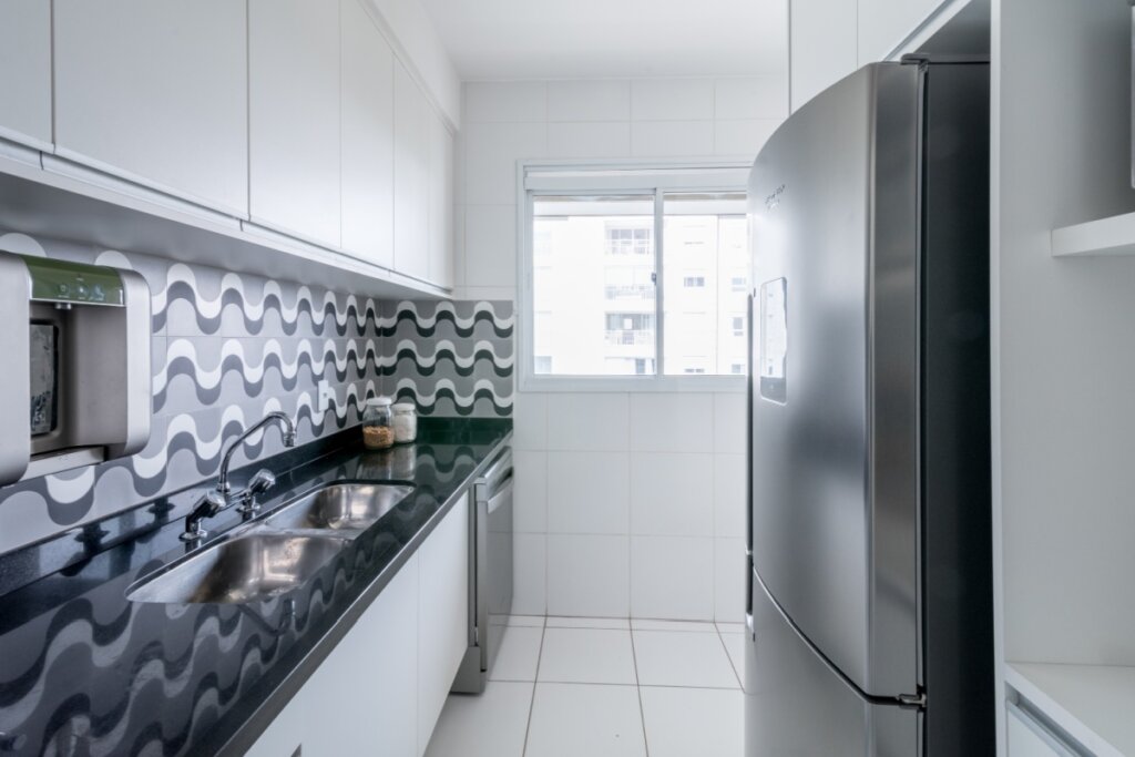 Cozinha de corredor (estilo galpão) com bancada de granito preto e cuba dupla de inox. O destaque é o revestimento da parede com padrão de ondas em tons de cinza, branco e preto. Armários aéreos brancos e uma geladeira de inox completam o ambiente compacto.