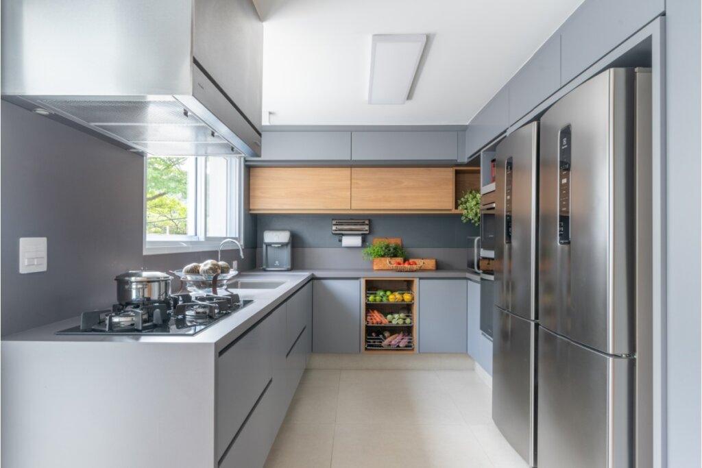 Cozinha moderna em formato de "U" com armários planejados em tom cinza fosco e detalhes em madeira clara. A bancada cinza integra um cooktop e uma cuba de inox sob a janela. Ao lado direito, duas geladeiras de aço escovado estão embutidas na marcenaria.