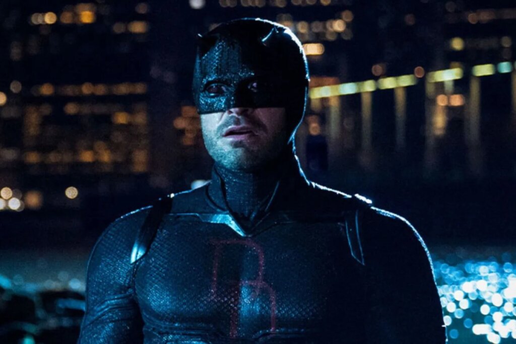 Cena noturna do herói Demolidor em um ambiente urbano. Ele está de frente, vestindo seu uniforme tático escuro com texturas que lembram escamas e o icônico logotipo "DD" vermelho no peito. O herói usa sua máscara característica e tem uma expressão de alerta ou surpresa, com a boca levemente aberta. Ao fundo, luzes de uma cidade e o reflexo da água criam pontos de brilho desfocados.