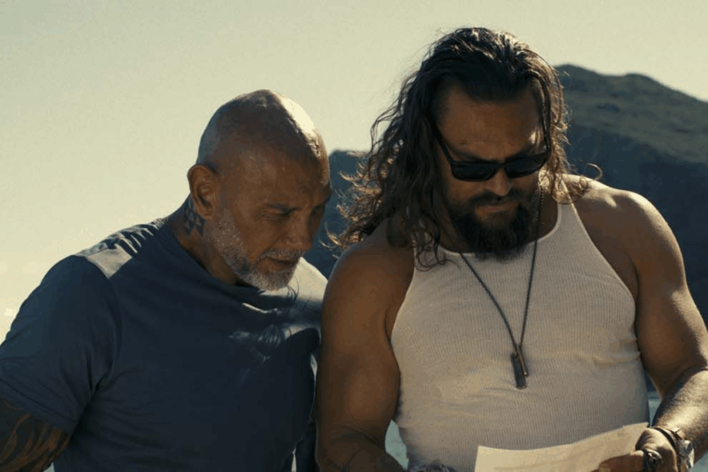 Dave Bautista e Jason Momoa aparecem lado a lado em um plano fechado, ambos olhando para baixo para um papel que seguram. Bautista, à esquerda, é careca com barba grisalha e usa camiseta azul; Momoa, à direita, tem cabelos longos, óculos escuros e usa uma regata branca. O fundo mostra uma paisagem litorânea ensolarada.