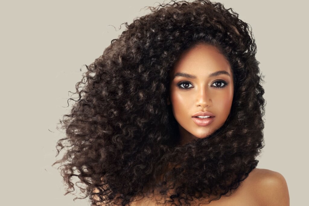 Mulher jovem com cabelos longos, volumosos e cacheados em tom castanho escuro, com brilho natural, olhando para a câmera sobre fundo neutro claro