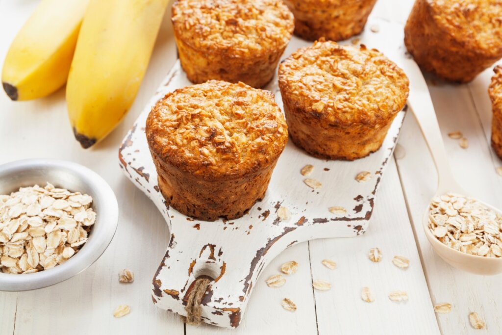 Três muffins de banana e aveia dourados sobre uma pequena tábua de corte branca e desgastada. Ao lado, duas bananas inteiras, uma tigela prateada com aveia em flocos e uma colher de madeira também cheia de aveia. Os itens estão sob uma mesa branca de madeira.