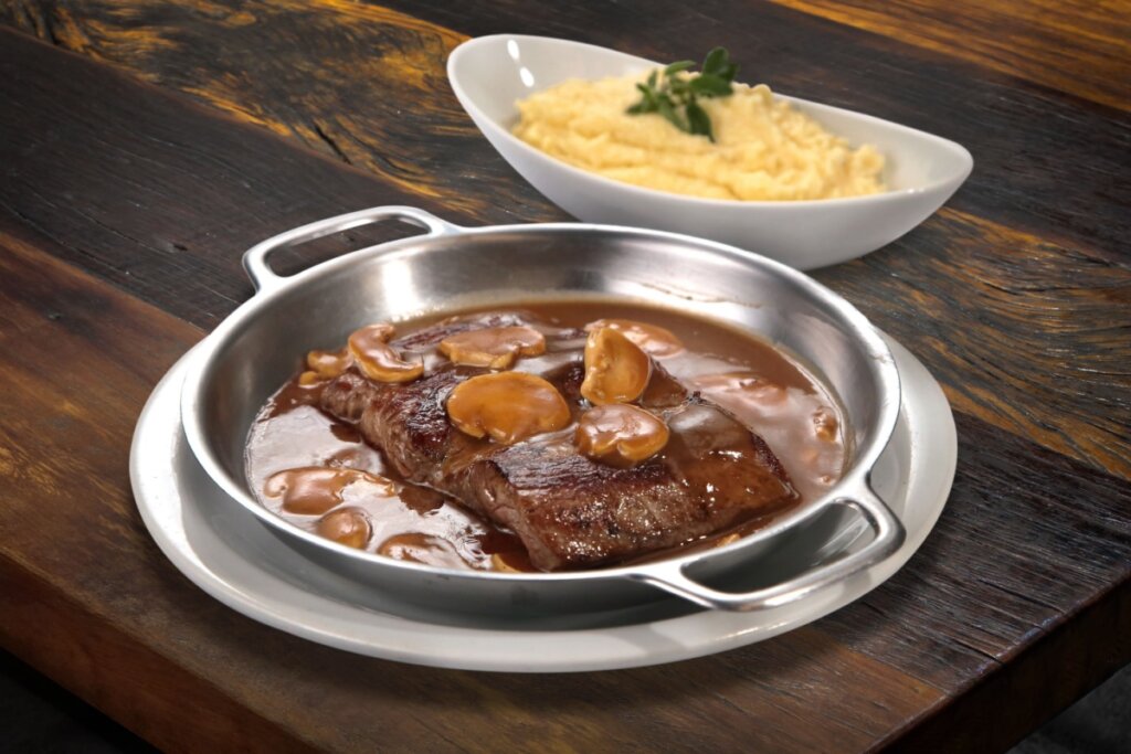 Panela de metal em cima de um prato branco com músculo ao molho madeira com cogumelos