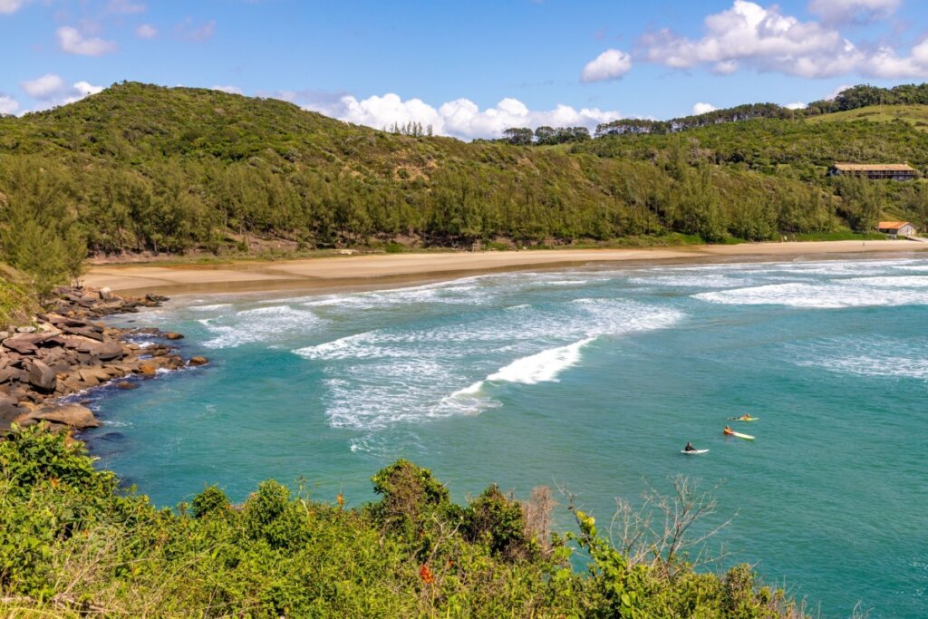 Vista aérea da Praia Vermelha, em Imbituba, com foco em uma enseada de águas claras em tons de turquesa e verde. No mar, três surfistas aguardam ondas em suas pranchas. A areia clara é emoldurada por encostas íngremes cobertas por densa vegetação nativa e árvores. No canto esquerdo, rochas escuras encontram a água, enquanto ao fundo, sob um céu azul com nuvens brancas.
