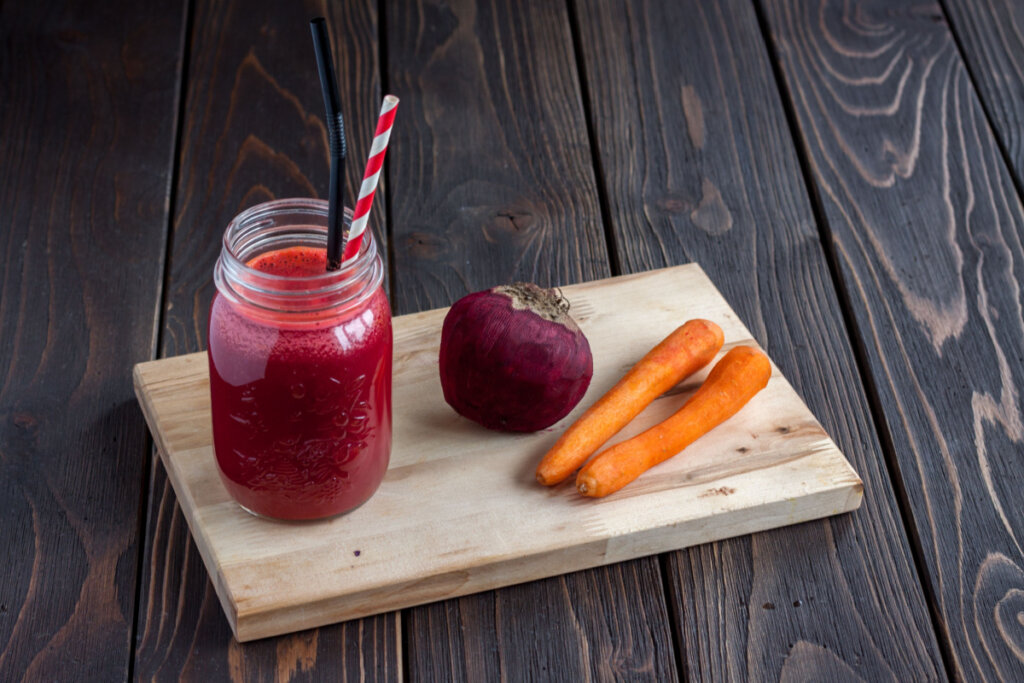 Foto de um suco de beterraba com cenoura, com duas cenouras e uma beterraba ao lado, em cima de uma tábua de madeira