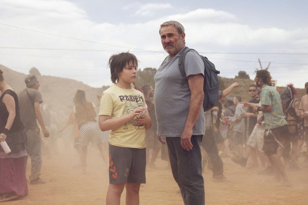 Cena do filme "Sirât" com os atores Sergi López e Bruno Núñez em pé em uma festa no deserto  