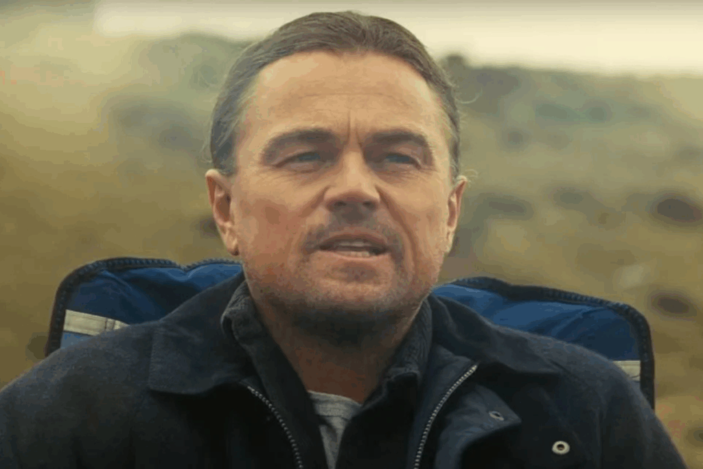 O ator Leonardo DiCaprio aparece em um plano médio, com uma expressão cansada e resoluta. Ele tem o cabelo escuro puxado para trás, uma barba rala e veste uma jaqueta azul-marinho sobre uma camisa cinza. Ele está sentado em uma cadeira de camping azul, com um fundo levemente desfocado que sugere um campo aberto sob uma luz amarelada e nebulosa, criando um clima introspectivo.