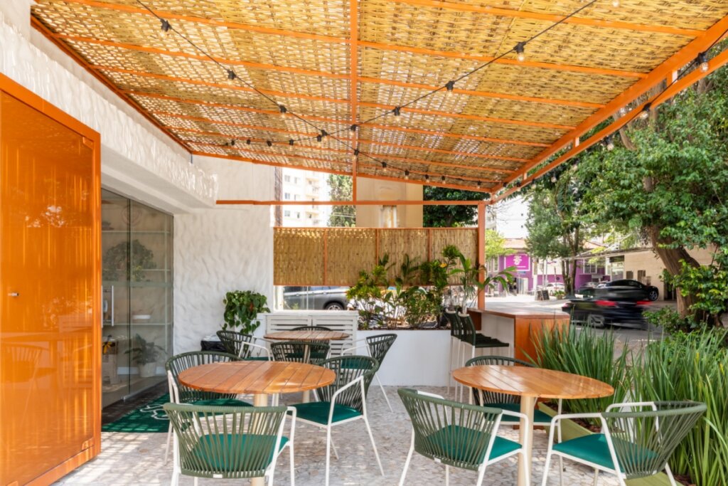 Área gourmet com cobertura artesanal, paredes brancas, mesas de madeira e cadeiras verdes