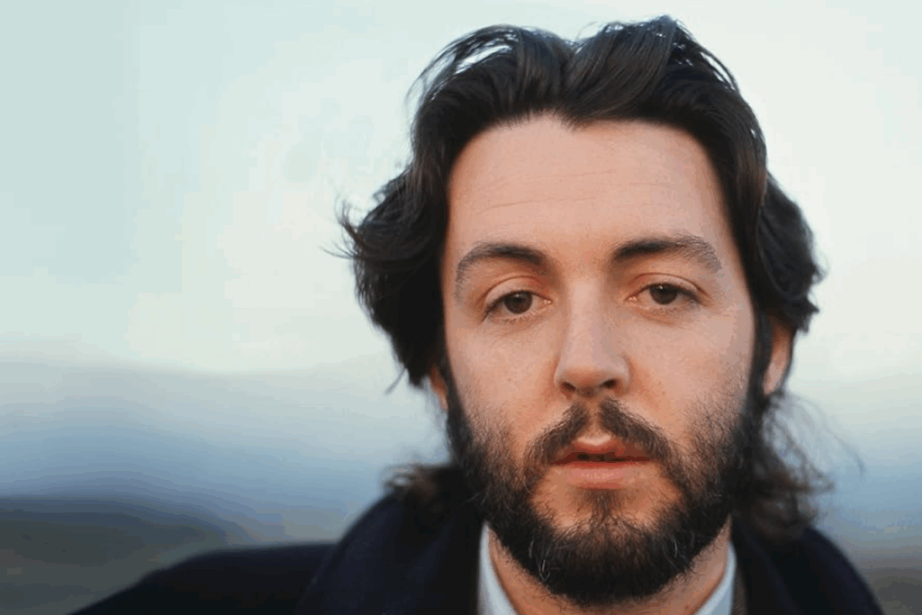 Homem com cabelo preto, curto, usando barba e camisa branca e casaco preto, olhando para frente em direção a câmera