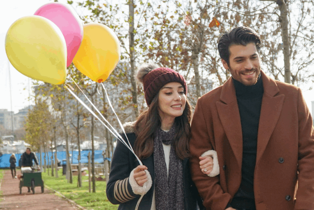 Casal andando no parque, o homem tem o cabelo curto e barba e está usando camisa preta e casaco marrom e a mulher está usando touca de frio vinho e cinza e casaco de frio preto com listras bege na manga e cachecol cinza e camisa branca. Com balões coloridos na mão