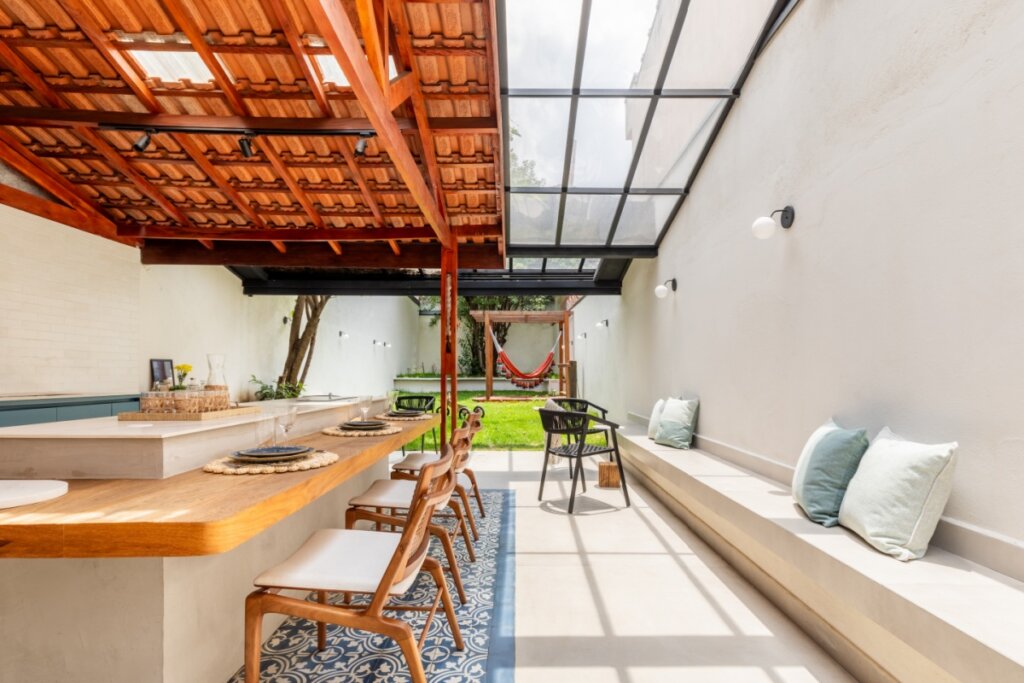 área externa com telhado de barro, telha translúcida e cobertura de vidro. Abaixo, uma bancada branca com cadeiras de madeira