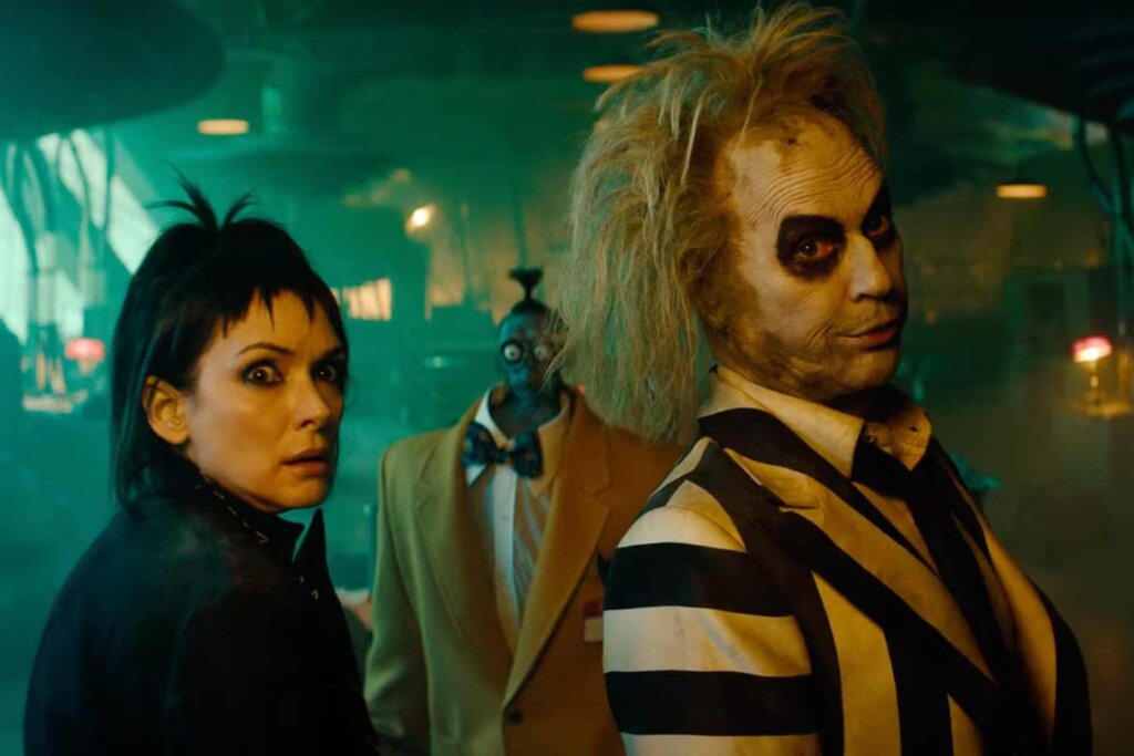 Cena do filme "Beetlejuice Beetlejuice" (2024). Em primeiro plano, à direita, está Beetlejuice (Michael Keaton), com seu icônico terno listrado de preto e branco, maquiagem cadavérica e cabelo verde bagunçado. À esquerda, Lydia Deetz (Winona Ryder) aparece com expressão de choque, usando roupas pretas e seu característico penteado com franja pontiaguda. Entre eles, ao fundo, vê-se um personagem com a cabeça encolhida usando um terno amarelo. O ambiente está imerso em uma iluminação esverdeada e enevoada, típica do "Mundo dos Mortos".
