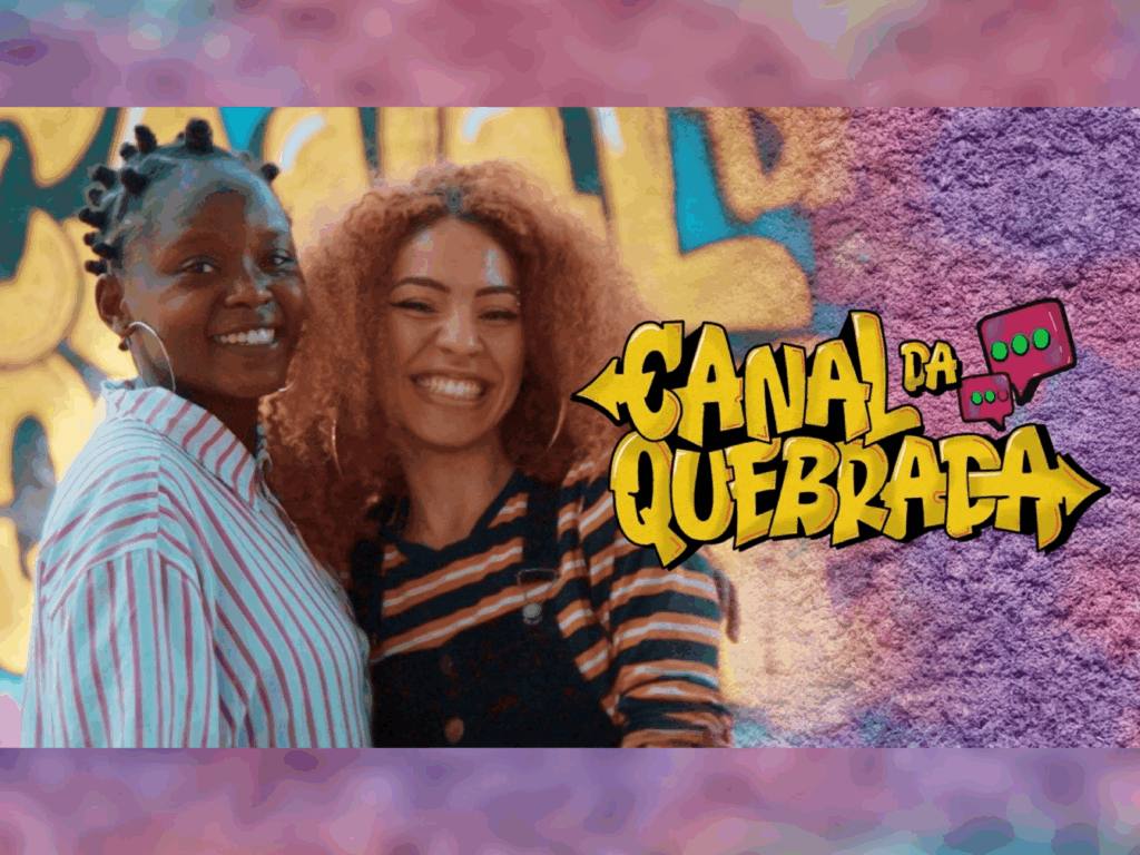 Arte promocional do "Canal da Quebrada". À esquerda, duas mulheres jovens sorriem alegremente; uma tem cabelos pretos presos em pequenos coques e veste camisa listrada, a outra tem cabelos cacheados ruivos e veste uma blusa listrada com macacão escuro. À direita, sobre um fundo de textura de parede urbana roxa, destaca-se o logotipo em estilo grafite amarelo com bordas pretas escrito "CANAL DA QUEBRADA", acompanhado de ícones de balões de conversa.