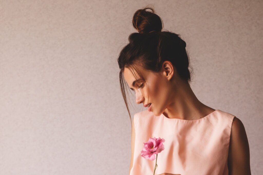 Fotografia de perfil de uma mulher jovem de pele clara, com o rosto levemente inclinado para baixo. Ela usa um coque alto e despojado no topo da cabeça, com duas mechas finas de cabelo soltas emoldurando o rosto. Veste uma blusa de cor rosa suave e segura uma flor rosa delicada à sua frente. O fundo é uma parede de textura simples em tom neutro, com iluminação suave vindo da lateral.