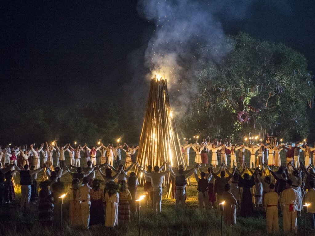 Cena noturna da série "Desalma" mostrando uma celebração tradicional. No centro, há uma grande fogueira alta feita de troncos de madeira em formato de cone, com chamas e faíscas subindo ao céu. Dezenas de pessoas em trajes típicos ucranianos (camisas brancas e coletes) formam um grande círculo ao redor do fogo, de mãos dadas e braços erguidos. À direita, há uma grande árvore decorada com fitas coloridas. Várias tochas iluminam o campo ao redor do grupo.