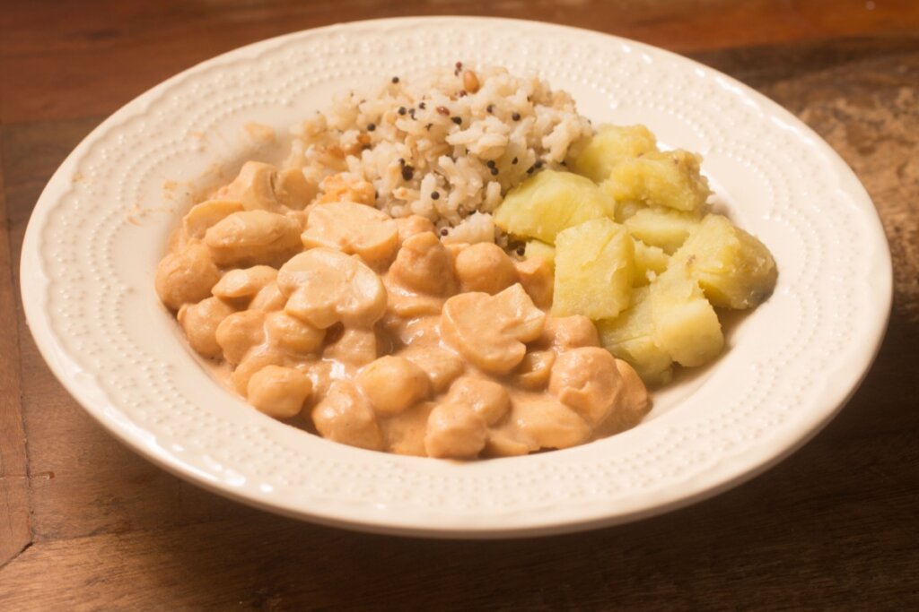 Um prato branco de cerâmica com bordas trabalhadas, servido com estrogonofe cremoso de grão-de-bico e cogumelos fatiados. O prato é acompanhado por uma porção de arroz integral com grãos mistos e uma porção de batatas cozidas picadas.