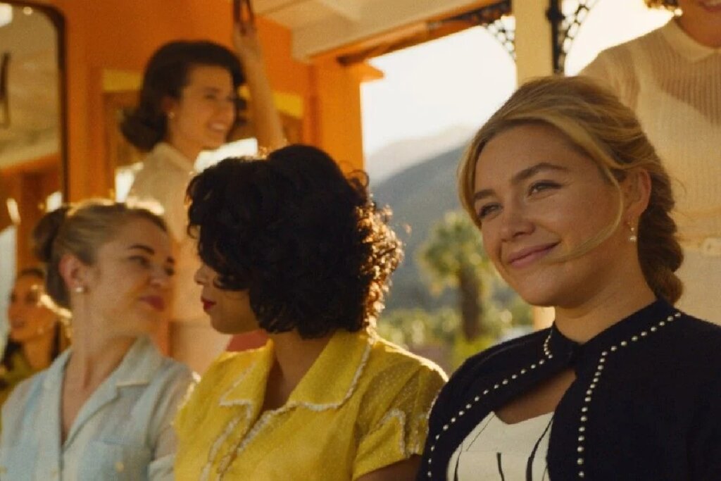 Close lateral da personagem de Florence Pugh no filme "Não Se Preocupe, Querida". Ela está sorrindo levemente, com o cabelo loiro preso e usando brincos de pérola. Ao fundo, outras mulheres com penteados e roupas no estilo dos anos 1950 aparecem em um ambiente ensolarado e em tons pastéis, sugerindo uma atmosfera utópica e retrô.