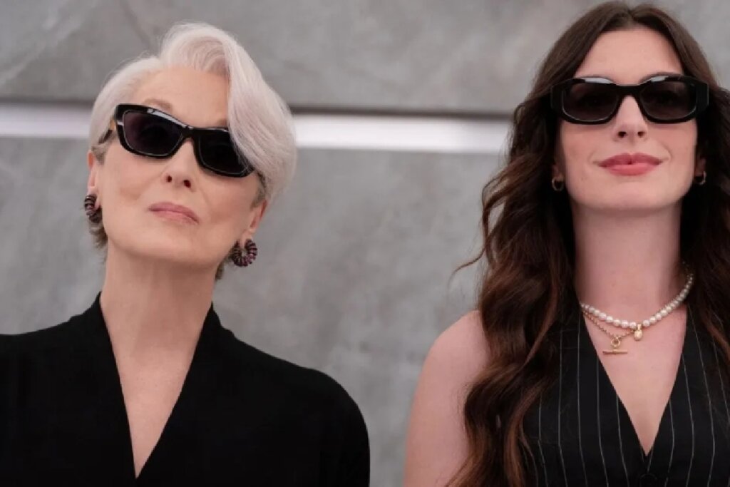 Plano médio das atrizes Meryl Streep e Anne Hathaway lado a lado. À esquerda, Meryl Streep exibe cabelos brancos curtos e usa óculos de sol pretos e uma blusa preta. À direita, Anne Hathaway tem cabelos castanhos longos e ondulados, usa óculos de sol pretos, um colete risca-de-giz e um colar de pérolas. Ambas têm expressões imponentes e elegantes contra um fundo cinza neutro