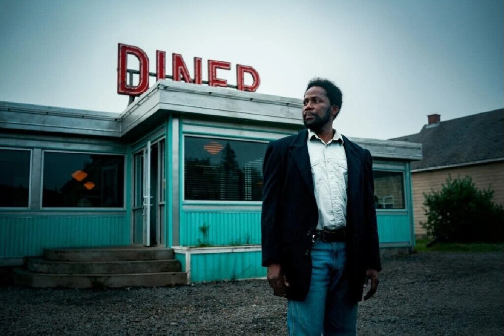 Imagem do ator Harold Perrineau como Boyd Stevens em cena da série "Origem", com expressão vigilante em um cenário nublado. À esquerda, destaca-se um restaurante retrô azul-turquesa com um letreiro em neon vermelho escrito "DINER".