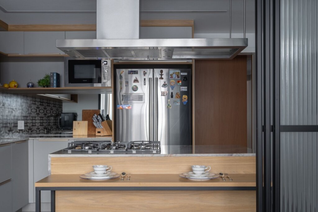 Esta imagem de uma cozinha moderna apresenta um piso de tábuas de madeira. A bancada da cozinha e a ilha de refeições apresentam superfícies de madeira de tábuas horizontais. O rack de vinho e as prateleiras abertas acima da bancada são feitos de madeira e metal. Um painel de madeira com veios horizontais reveste uma das paredes laterais, criando uma sensação de calor e continuidade.