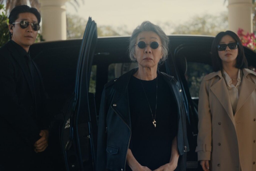 Três pessoas de expressão séria e óculos escuros posam à frente de um carro preto com a porta aberta. No centro, uma mulher idosa veste jaqueta de couro preta; à esquerda, um homem de terno preto; e à direita, uma mulher de sobretudo bege. O cenário é uma rua ensolarada com palmeiras ao fundo, evocando um clima de suspense cinematográfico.