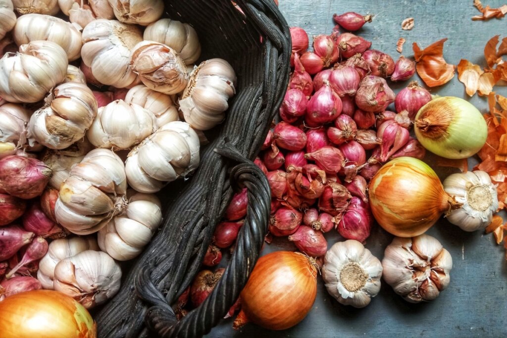 Alhos inteiros e descascados ao lado de cebolas espalhados sobre superfície de madeira, com uma cesta escura ao centro