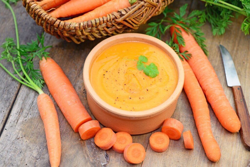 Creme de cenoura com cúrcuma servido em tigela de cerâmica em cima de mesa de madeira e com cenouras ao lado