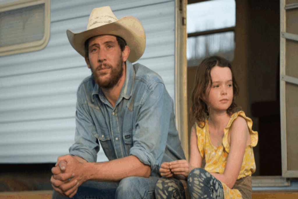 Homem com cabelo curto e barba usando chapéu branco e camisa e calça jeans com menina sentada ao lado com o cabelo curto solto, usando regata amarela e calça jeans com casa ao fundo