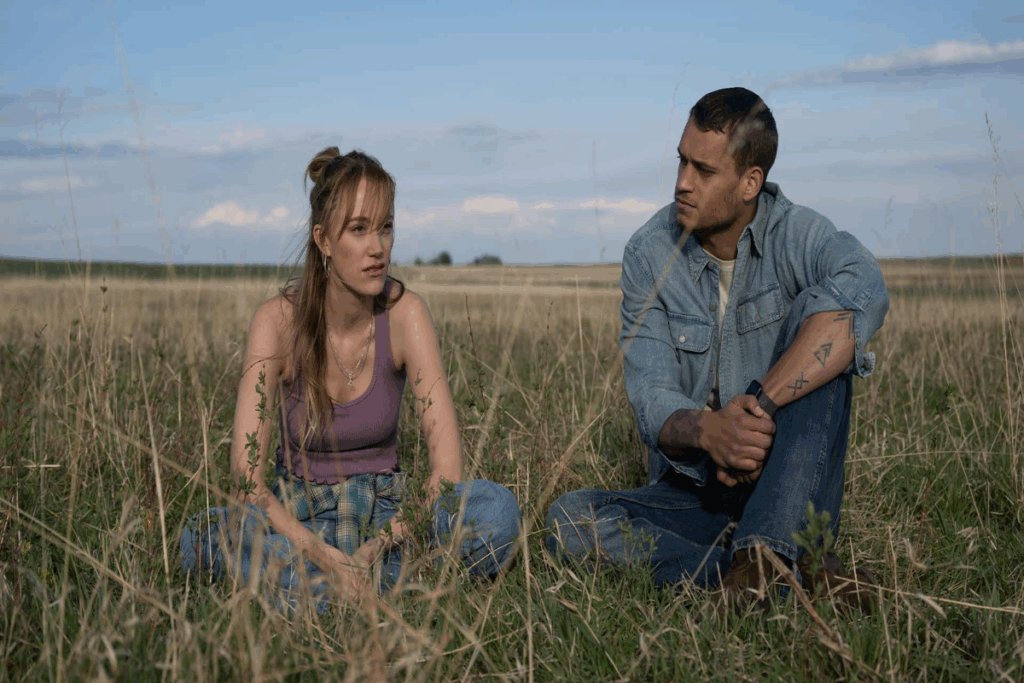 Mulher com cabelo preso parcialmente, usando regata roxa e calça jeans sentada em local aberto com grama ao lado de homem com cabelo curto e usando camisa jeans e calça jeans