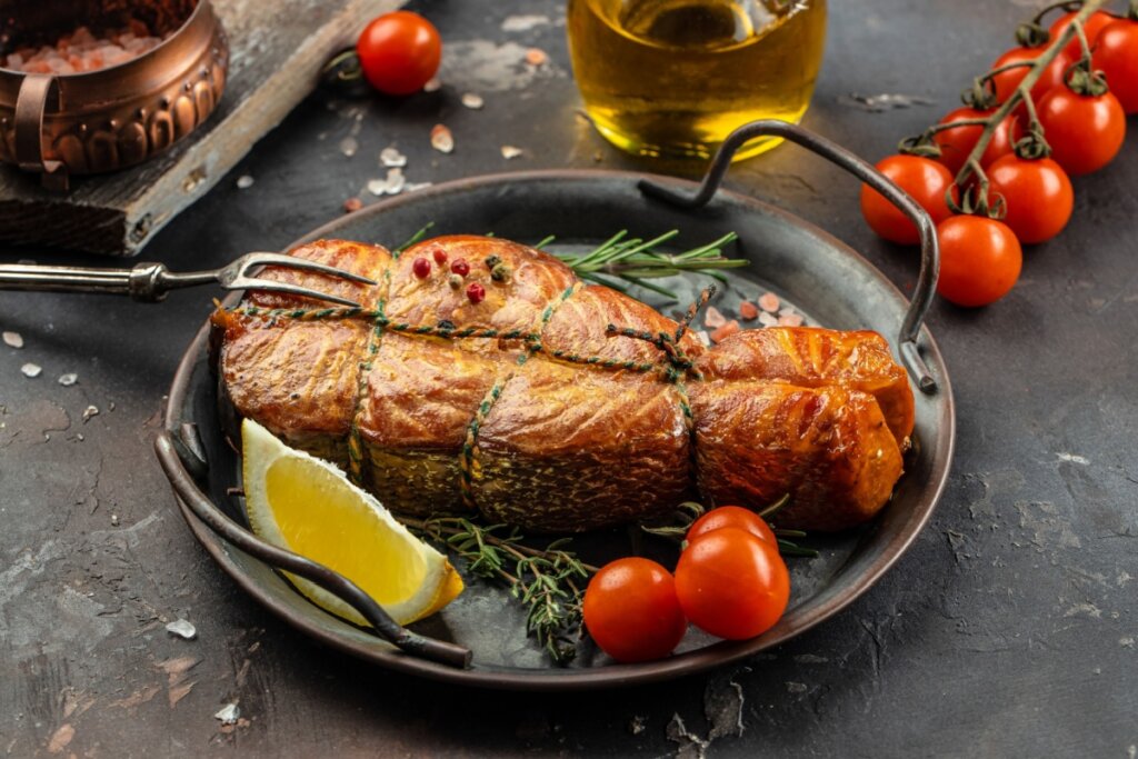 Esta imagem apresenta um suculento pedaço de salmão assado ou defumado, com uma textura dourada e brilhante, amarrado com barbante de cozinha para manter sua forma. O peixe está acomodado em uma frigideira de metal rústica, decorado com grãos de pimenta rosa, ramos de alecrim e tomilho fresco, além de tomates-cereja vibrantes e uma fatia de limão siciliano.