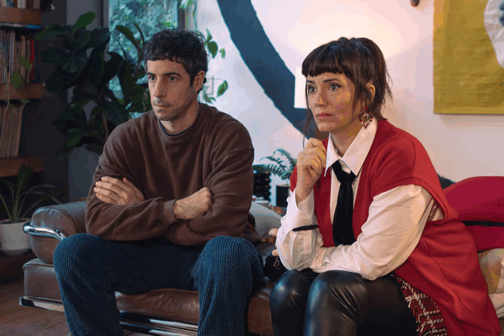 Casal sentado em sofá, o homem tem o cabelo preto e está usando moletom marrom e calça jeans e a mulher tem o cabelo preso e franja, usando camisa branca, gravata preta, colete vermelho e calça de couro preta