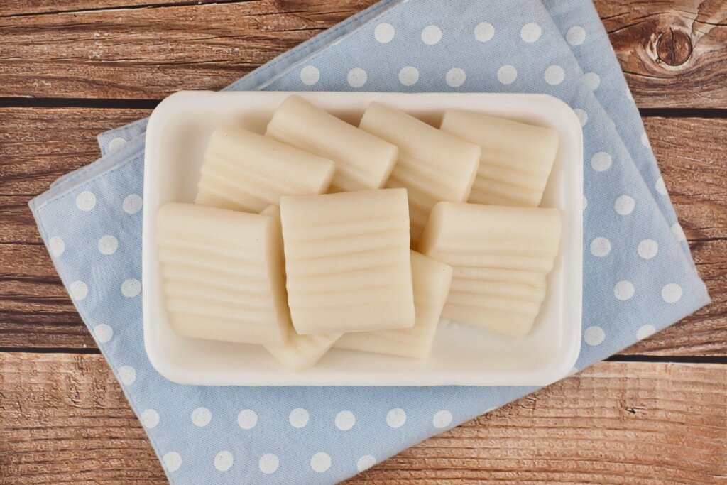 Vista superior de uma bandeja branca de isopor contendo várias unidades de Jeolpyeon, um bolo de arroz tradicional coreano. Os bolinhos são de cor branca, possuem formato retangular e apresentam ranhuras lineares paralelas em sua superfície, feitas por carimbos tradicionais. A bandeja está posicionada sobre um pano azul claro com estampa de bolinhas brancas, que por sua vez descansa em uma superfície de madeira rústica.