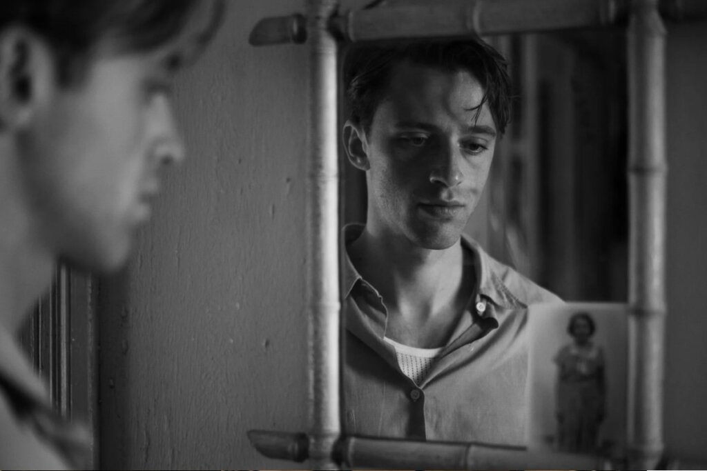 Imagem em preto e branco mostrando um homem jovem refletido em um espelho com moldura de bambu. O reflexo foca em seu rosto, que demonstra uma expressão melancólica ou pensativa. No canto inferior direito do espelho, está presa uma pequena fotografia antiga de uma mulher. O rosto real do homem aparece em primeiro plano, desfocado, à esquerda.