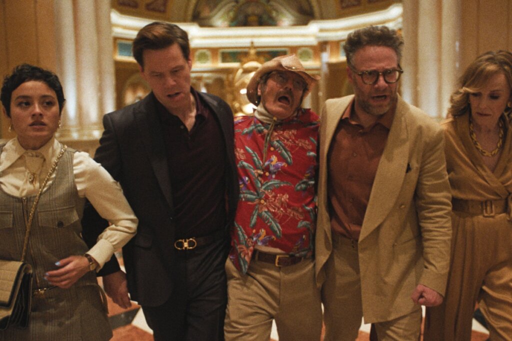 Fotografia de cena da série "The Studio". No centro, um grupo de cinco pessoas caminha apressadamente em um ambiente luxuoso de cassino ou hotel. Dois homens carregam um terceiro homem idoso, que usa uma camisa floral vermelha e chapéu de cowboy, e parece estar passando mal ou desmaiado. À esquerda, uma mulher jovem com expressão preocupada e, à direita, uma mulher loira com um macacão dourado observa a cena.