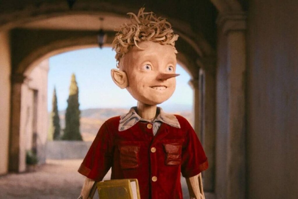 Close-up do boneco Pinóquio em uma animação. Ele é feito de madeira clara com texturas visíveis, tem olhos azuis expressivos e um sorriso leve. Ele veste uma camisa de veludo vermelho e carrega um livro amarelo sob o braço. O fundo mostra um arco de pedra abrindo-se para uma paisagem ensolarada com árvores.