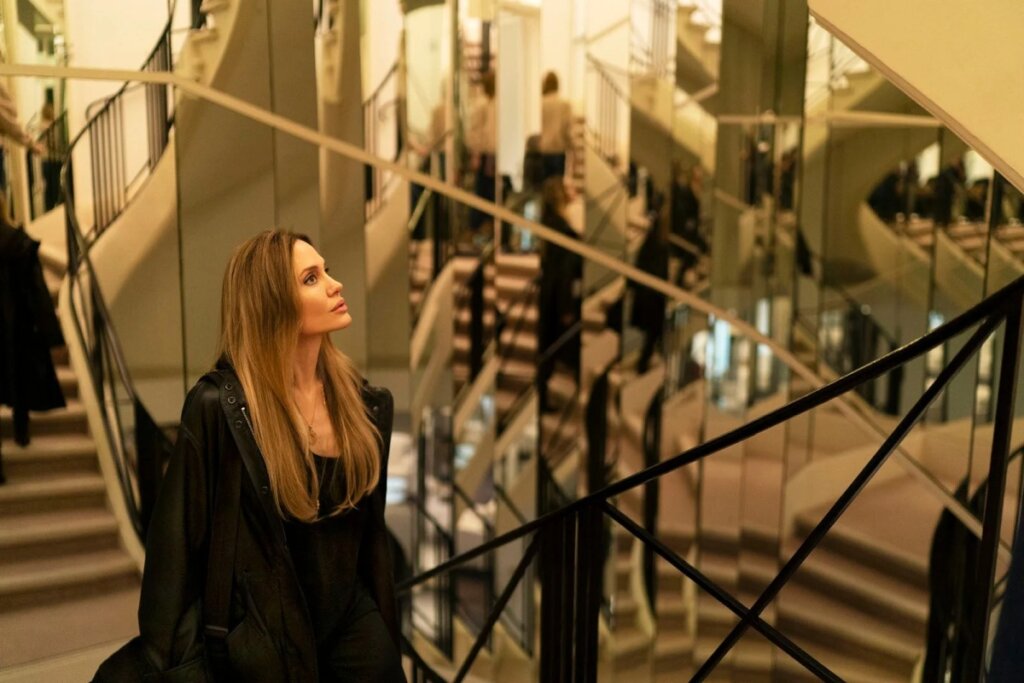 Fotografia de Angelina Jolie em um ambiente interno luxuoso com uma escadaria monumental ao fundo. Ela está de perfil, olhando para cima com uma expressão serena. O cenário é marcado por múltiplos espelhos que refletem a escada e sua própria silhueta, criando um efeito visual complexo de repetição e profundidade. Ela veste um casaco preto longo.