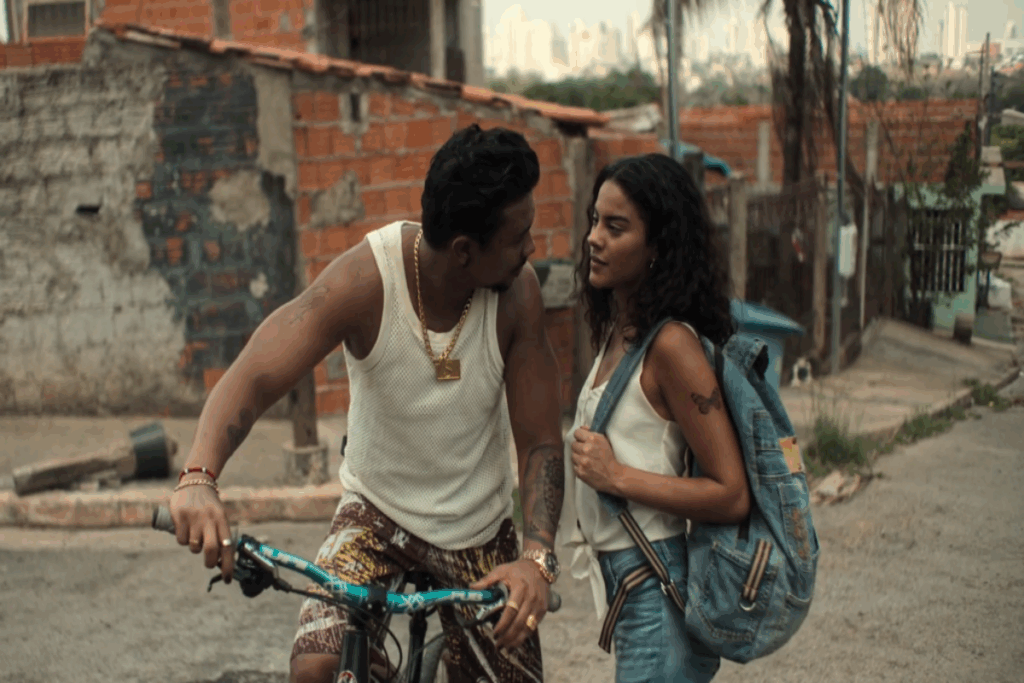 Mulher com cabelo solto, cacheado usando regata branca, calça jeans e com mochila nas costas azul conversando com homem que está em bicicleta com o cabelo curto, usando cordão dourado, regata branca e short estampado