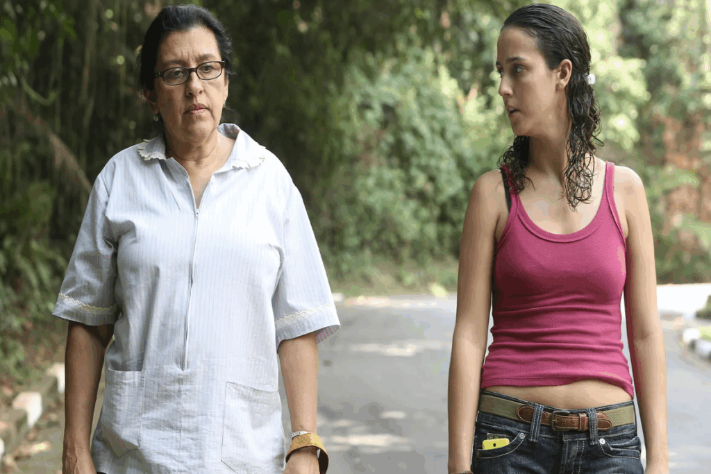 Imagem de uma mulher mais velha com cabelo preso, usando óculos preto e camisa de manga curta branca caminhando ao lado de jovem com o cabelo curto cacheado preso parcialmente usando uma regata rosa e calça jeans