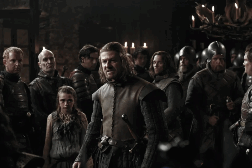 Homem de cabelos perto do ombro diante de um grupo em ambiente medieval na série Game of Thrones