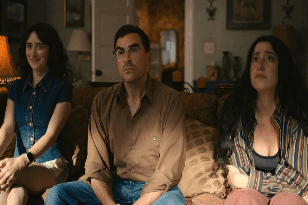 Duas mulheres e um homem sentados no sofá, uma das mulheres tem o cabelo solto, está usando uma regata jeans e short branco e está sorrindo, o homem está usando uma camisa de manga longa bege, calça jeans e óculos preto e tem o cabelo curto, e a outra mulher está usando uma calça rosa, blusa listrada e regata preta e está com o cabelo solto 