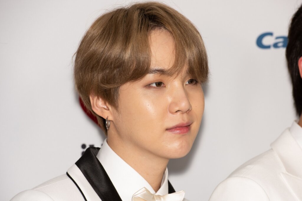 Homem com cabelo castanho claro partido ao meio com franja, usando camisa branca  e terno bege com detalhes em preto e gravata borboleta bege