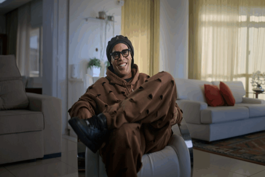 Jogador de futebol Ronaldinho Gaúcho, usando roupas de moletom marrom com rasgos sentado em cadeira em sala de estar sorrindo e usando óculos de sol e touca preta